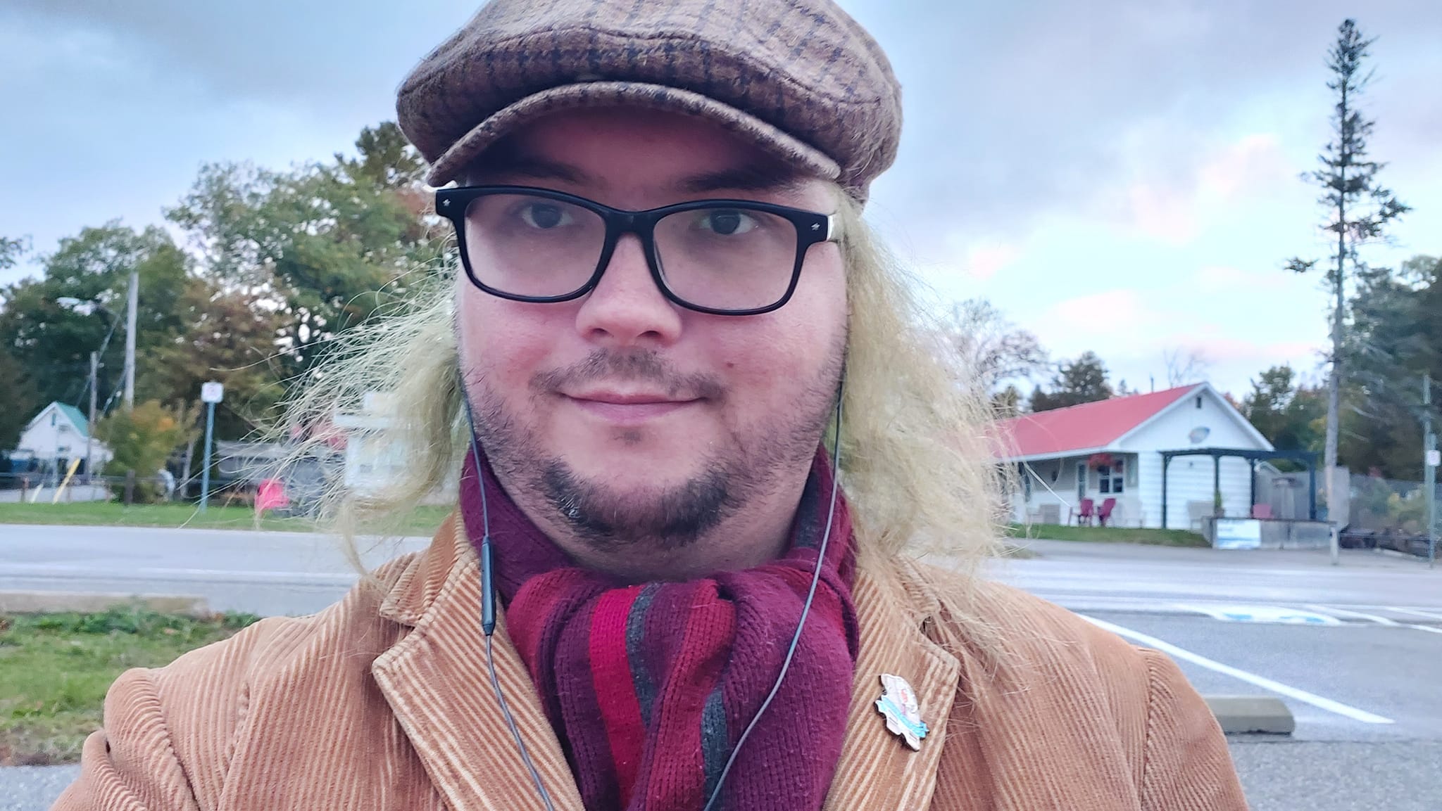 A man with long green hair stands on a sidewalk, a lakeside motel behind him